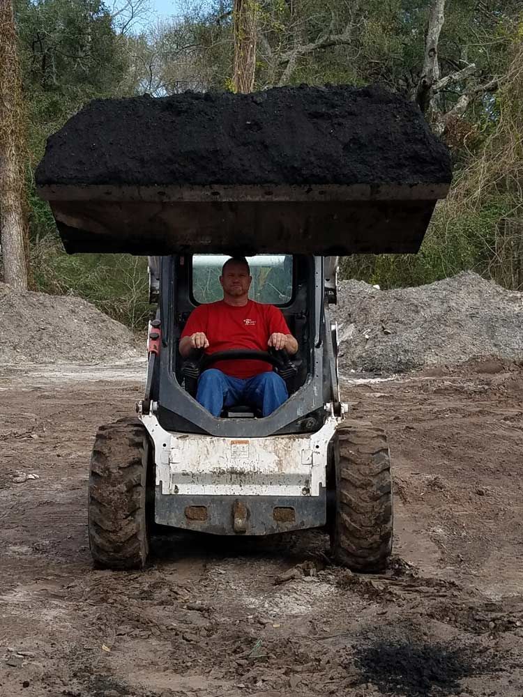 Skid steer loader carrying a large pile of soil on a muddy dirt path in a wooded area