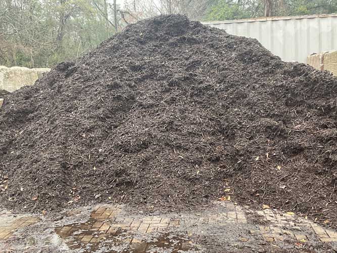 Large dark mulch or compost pile outdoors beside trees and a shipping container.