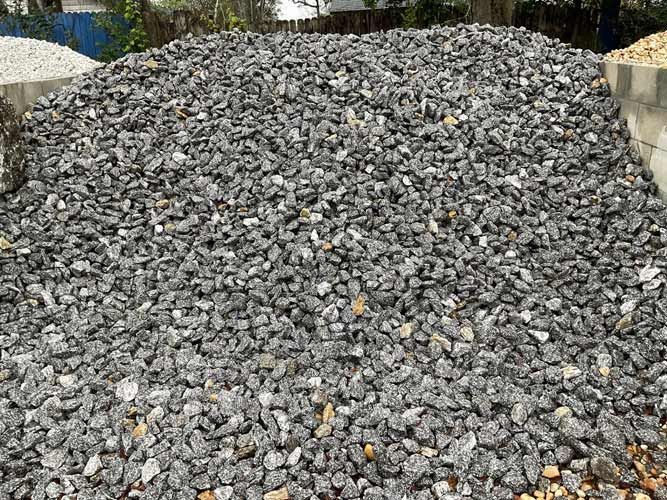 Large pile of gray gravel or crushed stone outdoors, with some greenery and concrete edges in the background