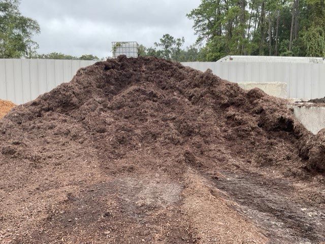 Large mound of dark soil in a yard, with a white fence and trees in the background