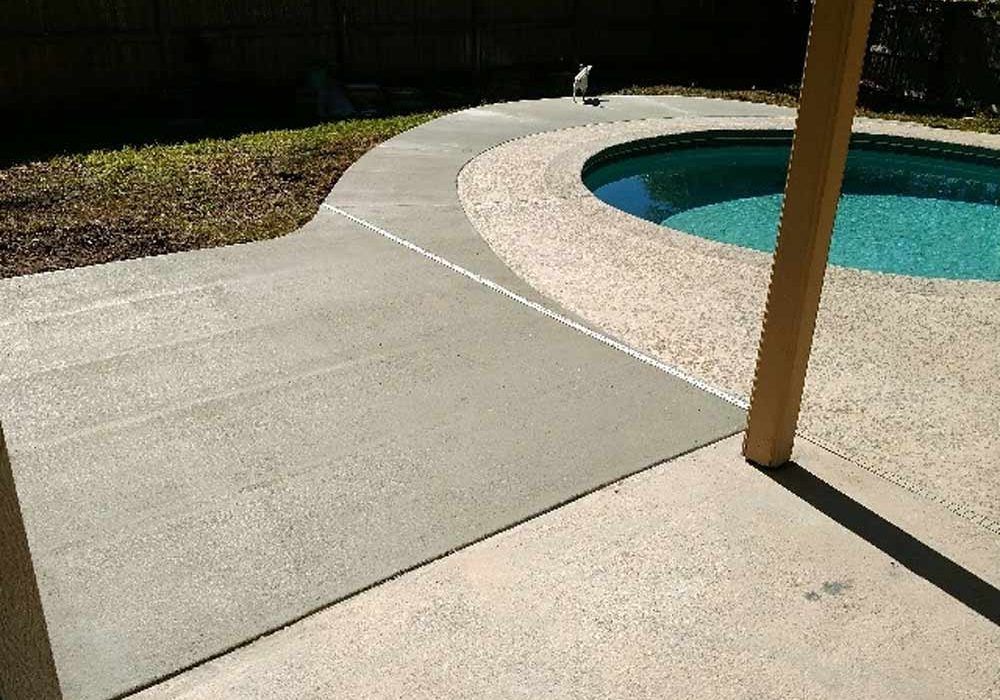 Concrete pool deck with curved walkway beside a blue swimming pool, shaded by a wooden post.