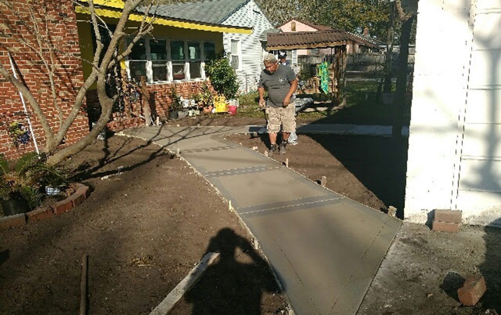 Person walking on a sunlit concrete path beside a brick house and yard greenery