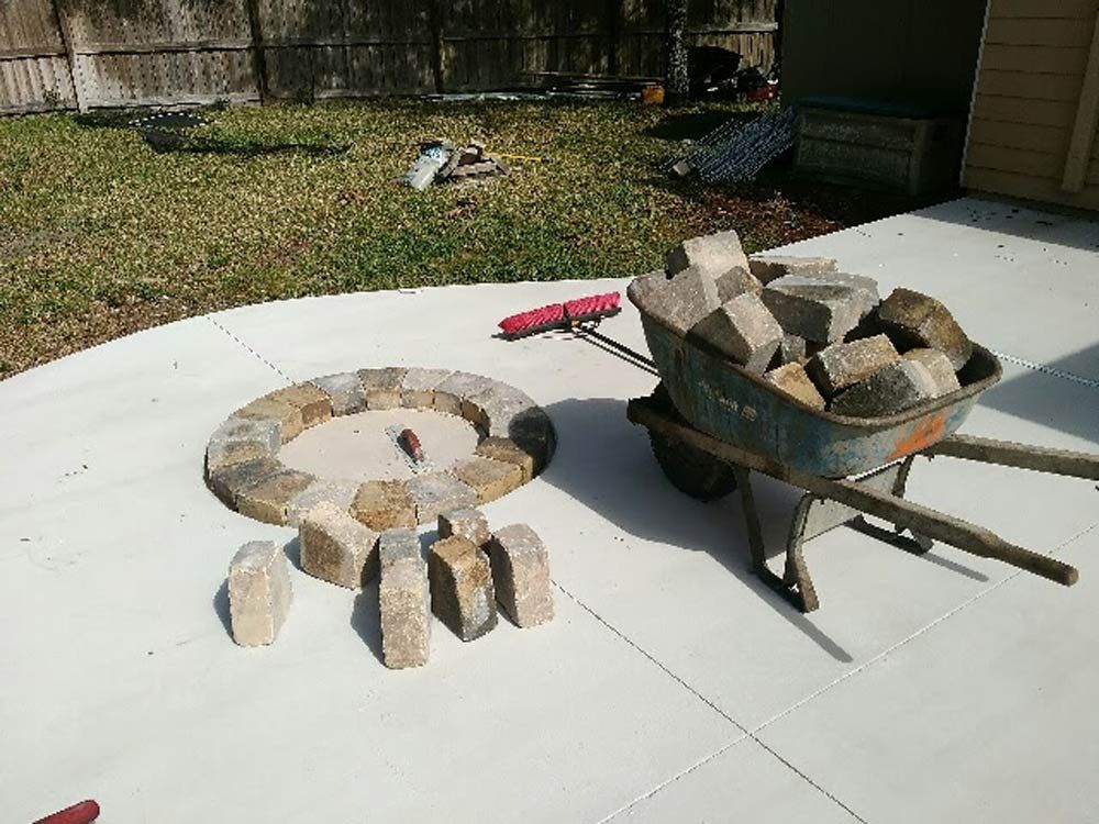 Metal fire pit and wheelbarrow filled with paving stones on a concrete patio in a backyard