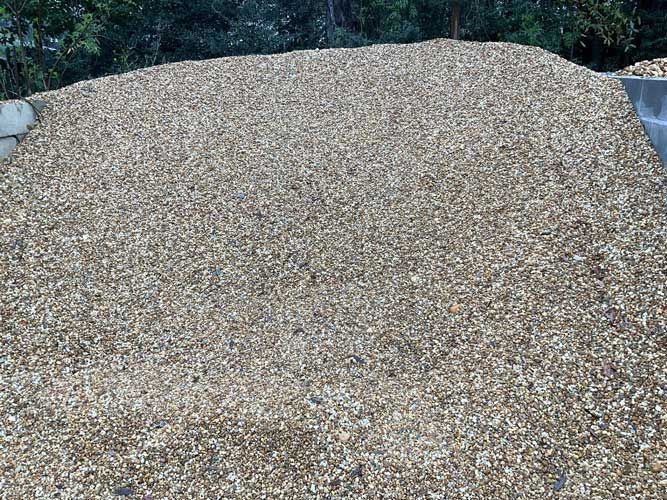 Large pile of small tan gravel or pebbles outdoors, with greenery and rocks at the edges