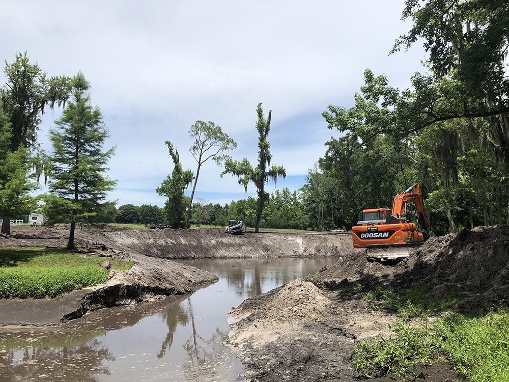 Orange excavator at muddy creek construction site with trees and cloudy sky