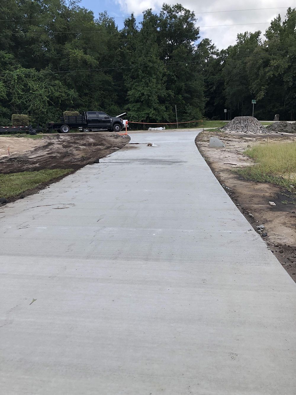 Concrete walkway under construction leading toward trees, with a pickup truck parked on the left and gravel on the right