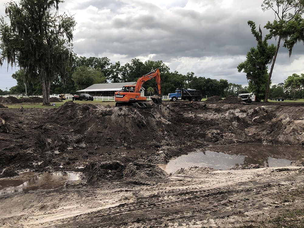 Excavator working in a muddy excavation site under cloudy skies, with trees and vehicles in the background