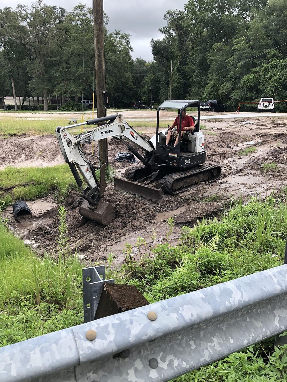 Small excavator digging muddy soil beside a road, with a guardrail and trees in the background.