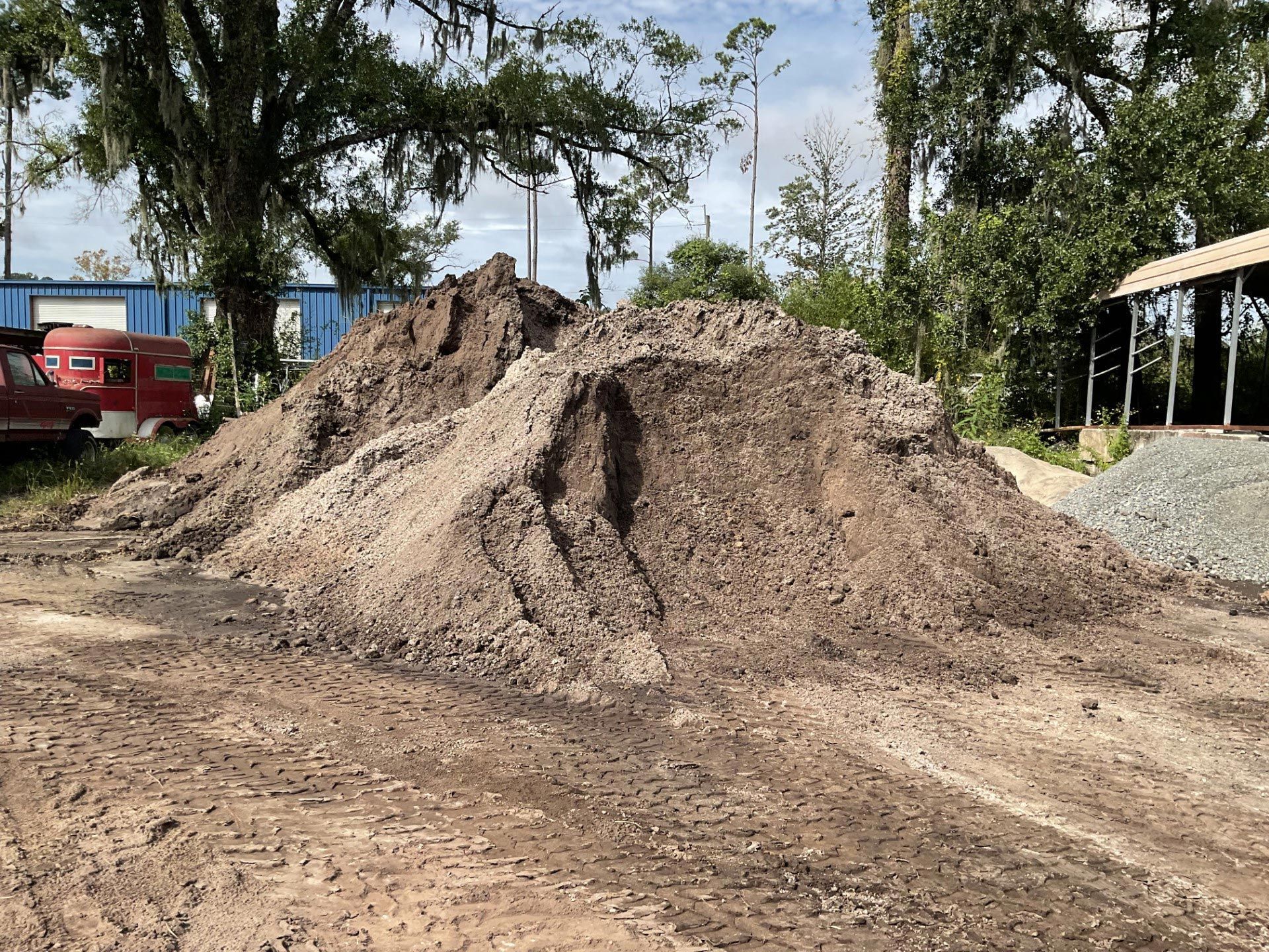 Large dirt pile on a construction site with trees, a red truck, and gravel in the background