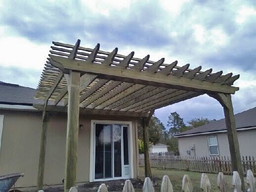 Backyard patio with a wooden pergola over sliding glass doors, beside a fenced yard under cloudy sky