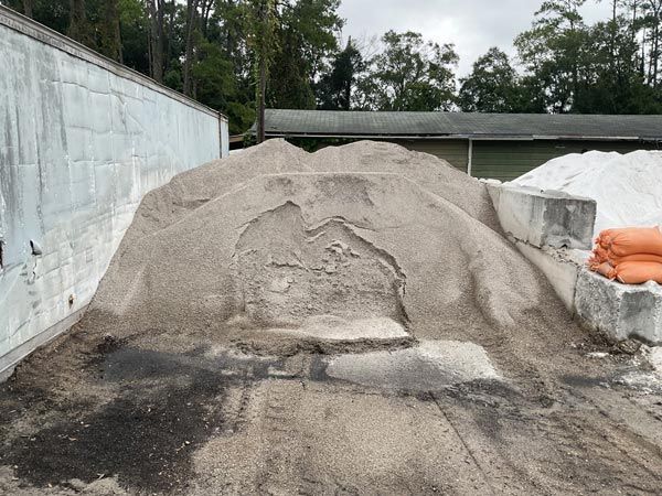 Large piles of gray gravel and sand in an outdoor storage yard beside a wall and orange sacks