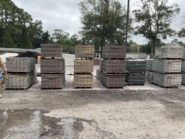Stacks of concrete pavers on pallets in an outdoor gravel lot with trees in the background