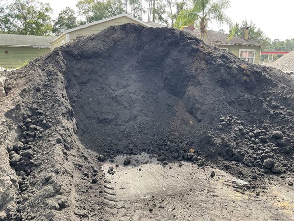 Large mound of dark soil or compost in a yard, with houses and trees in the background