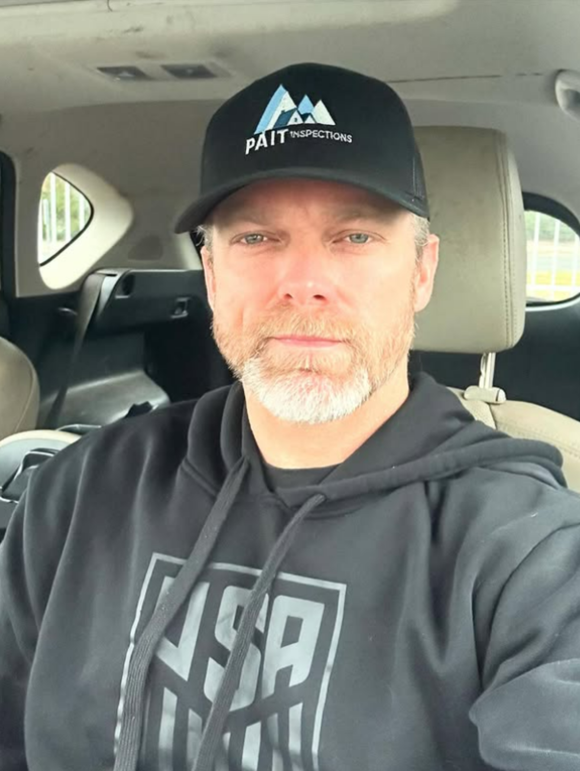Man in a black hoodie and hat, inside a vehicle, looking at the camera.