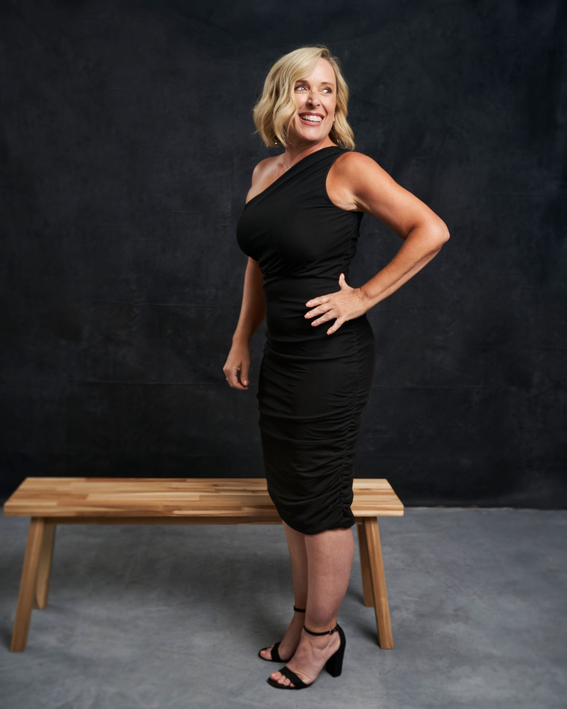 A woman in a black dress is standing next to a wooden bench.