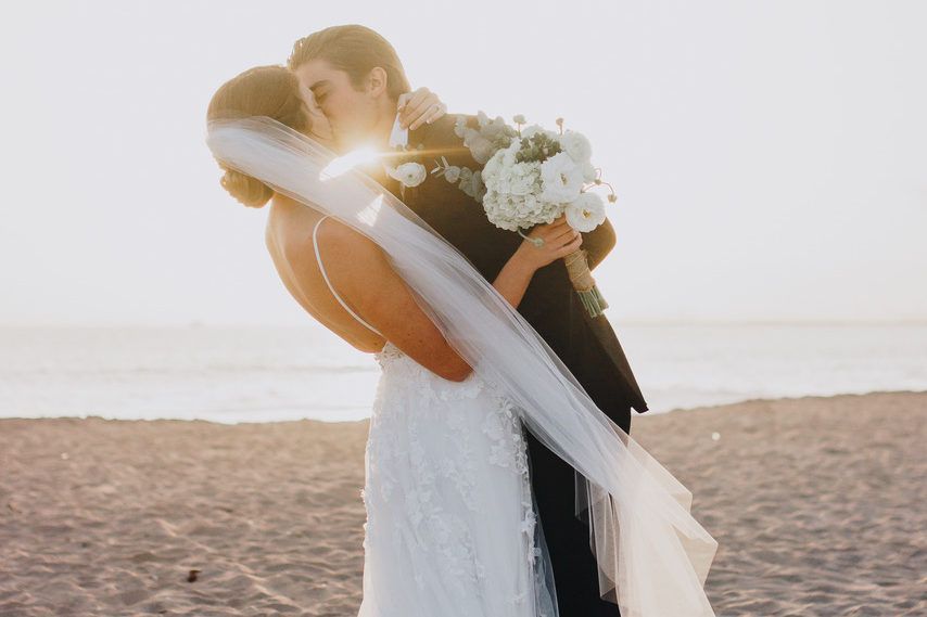 A bride and groom are kissing on the beach at sunset.