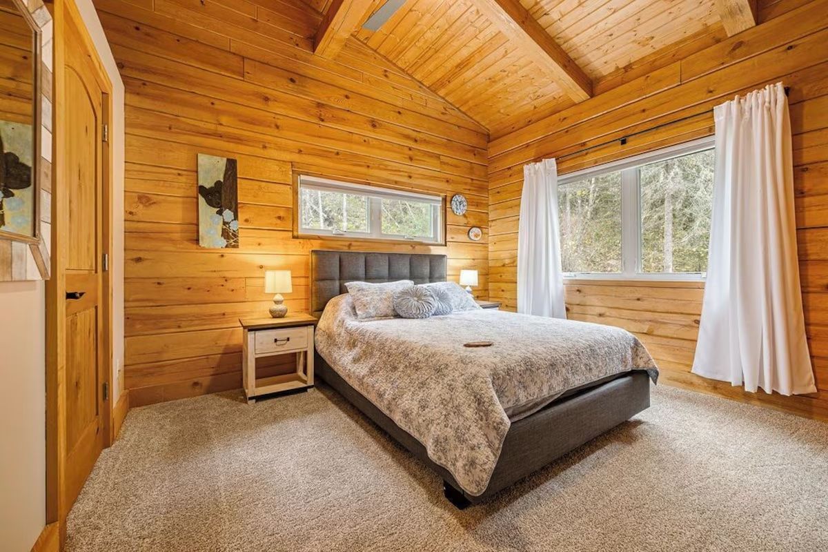 Cozy bedroom with wooden walls, bed, window, and curtains. Light-colored carpet.