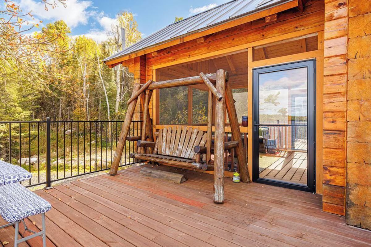Wooden porch with swing and glass door. Exterior of a cabin in the woods. Sunny, daytime.