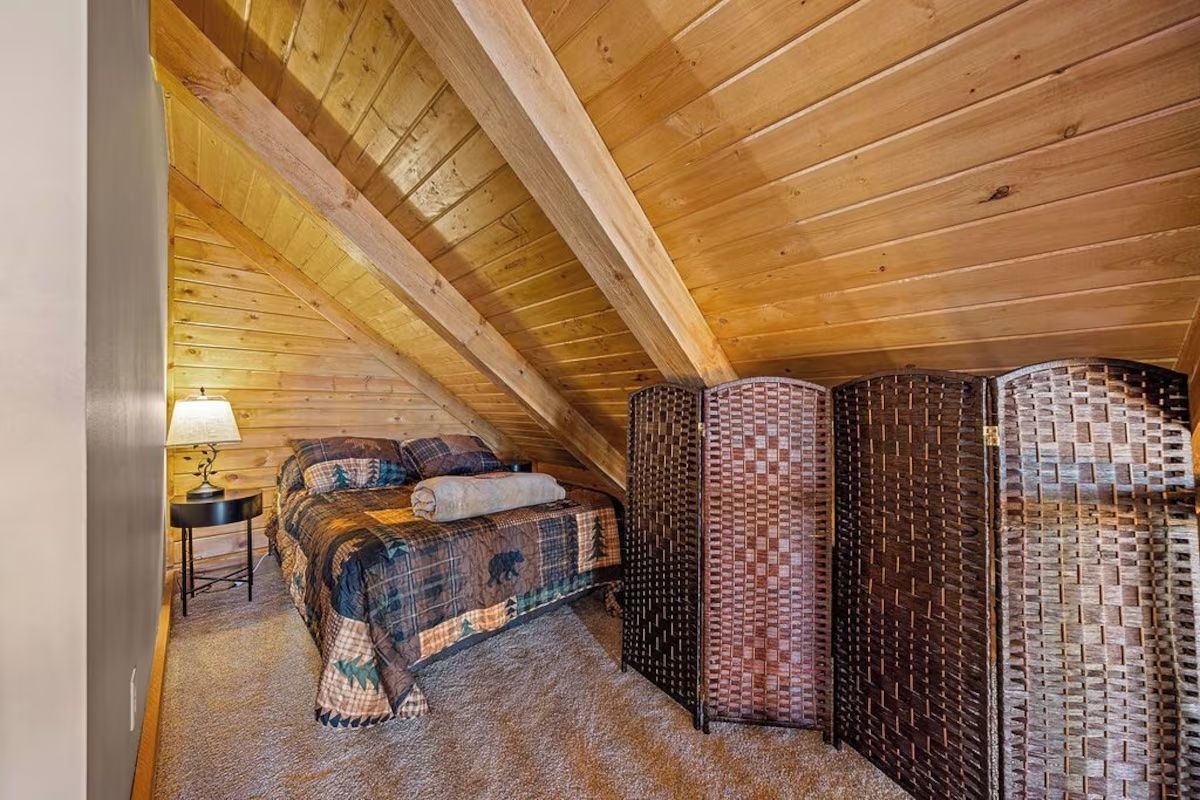 Cozy attic bedroom with sloped wood ceiling, bed, nightstand, screen, and carpet.