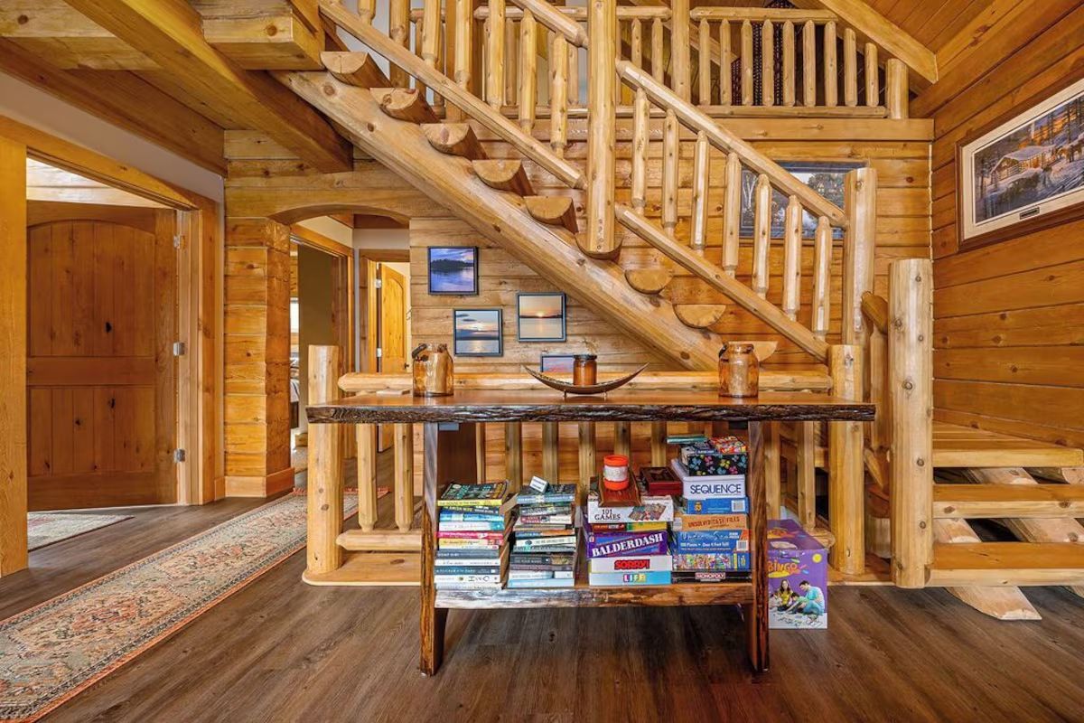 Rustic cabin interior with log staircase, wooden table with snacks, and framed artwork.