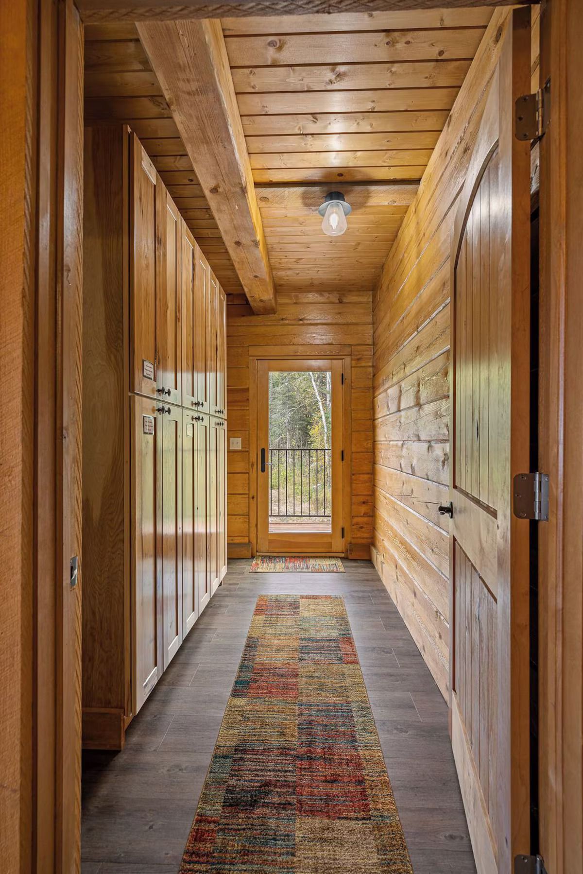Narrow wooden hallway with a colorful rug, wooden cabinets, and an exterior door.