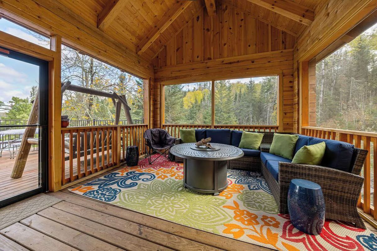 Screened-in porch with blue sectional, colorful rug, gas fire pit, and swing overlooking a forest.