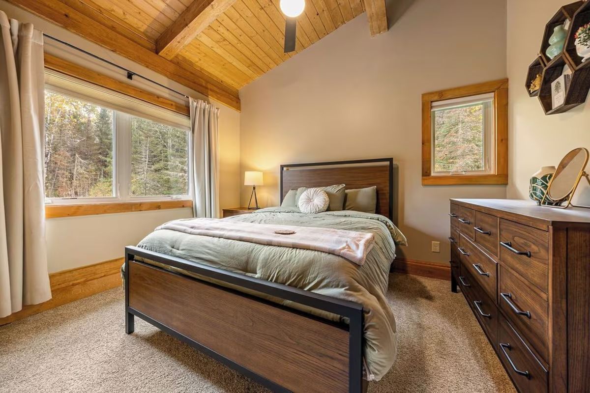 Bedroom with wooden bed frame, dresser, window overlooking trees, and light-colored walls.