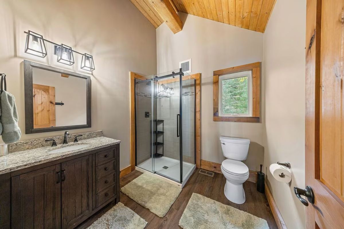 Bathroom with dark wood vanity, glass shower, and toilet; light beige walls and wood ceiling.