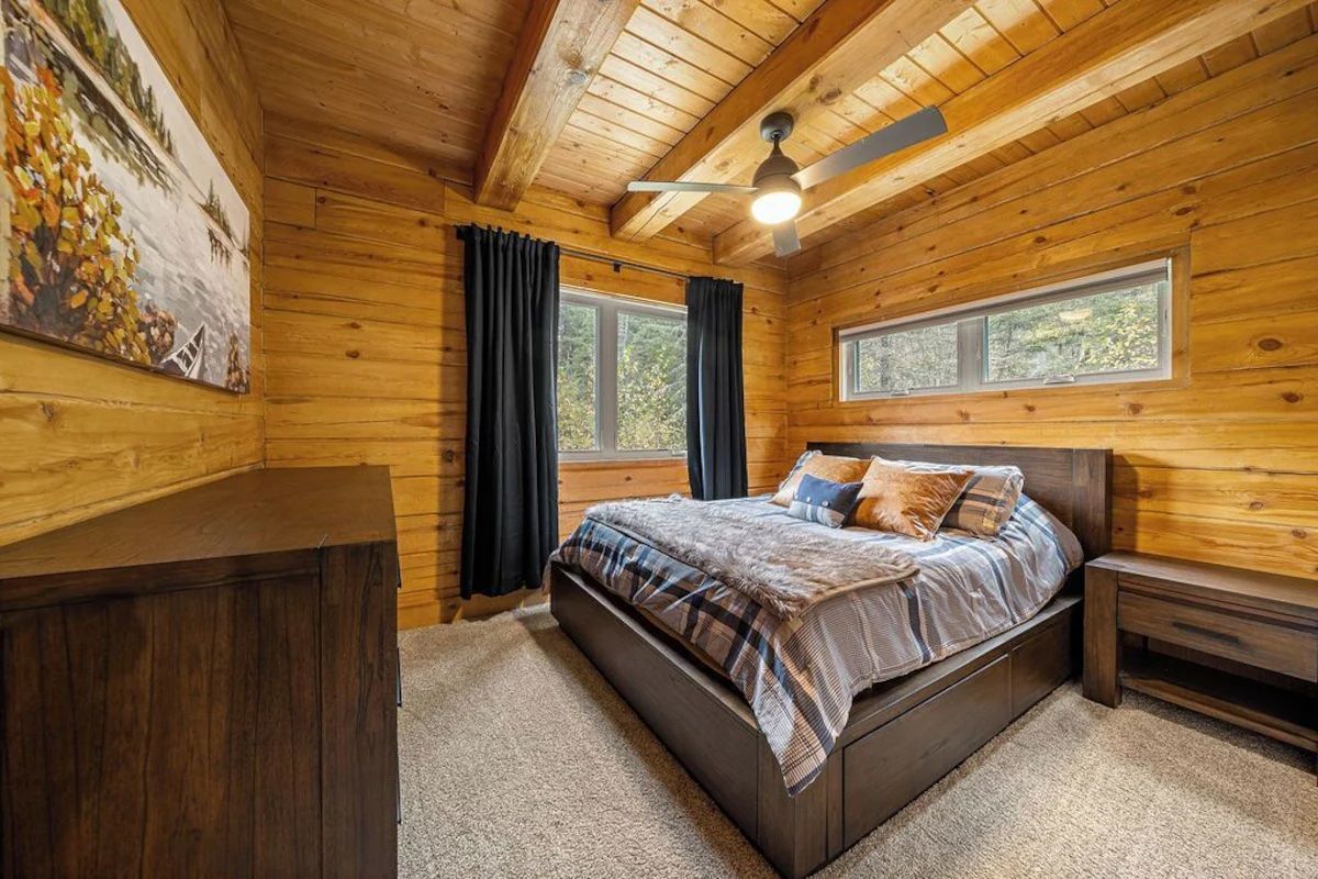 Bedroom with wood paneled walls and ceiling, bed, dresser, and window with dark curtains.
