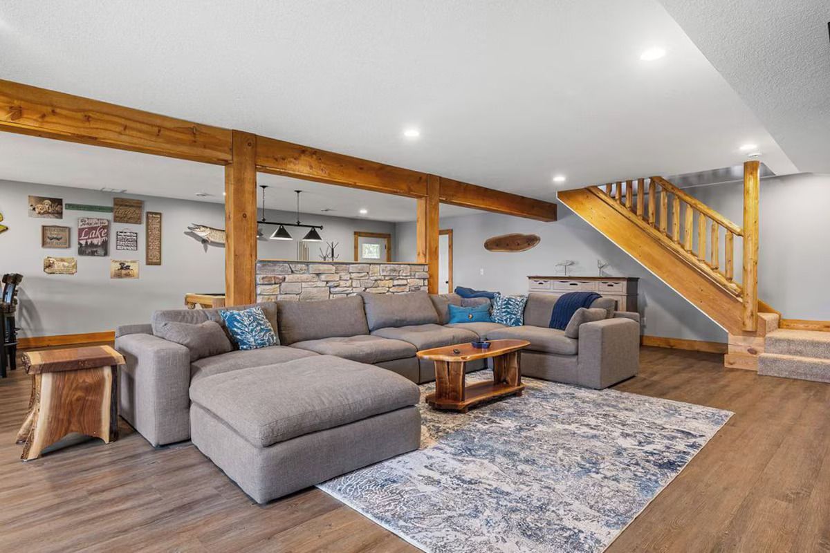 Cozy basement living room with gray sectional sofa, wood beams, and stairs.