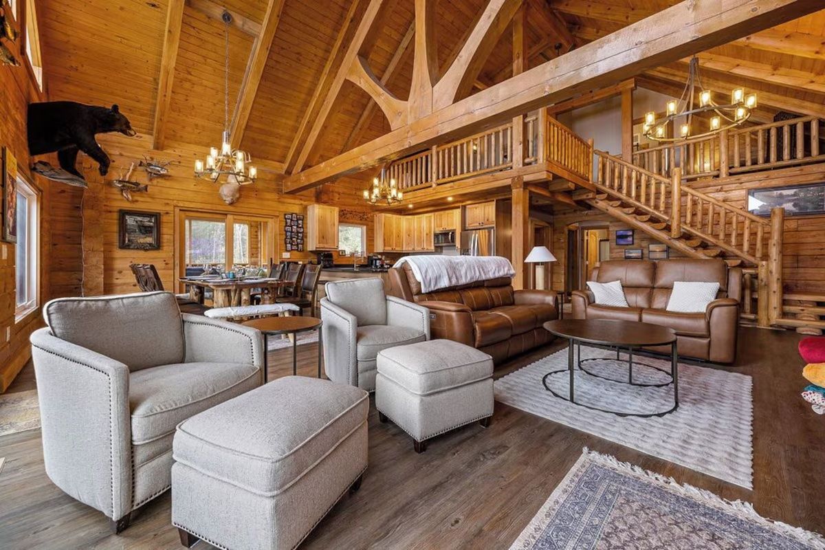 Rustic cabin interior with exposed wooden beams, multiple seating areas, and a staircase.