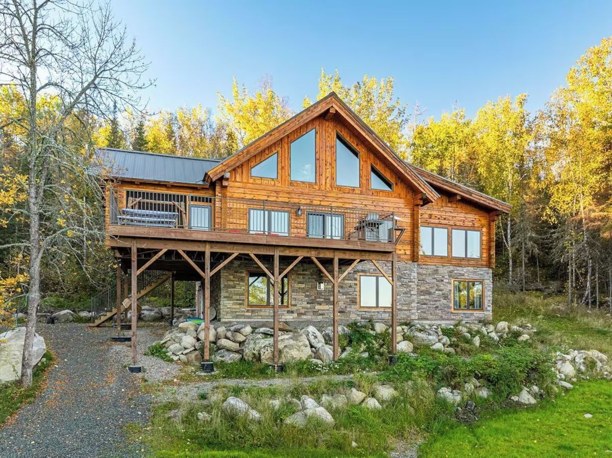 Two-story wood and stone cabin with a wraparound porch and tall windows, set among trees and rocks.