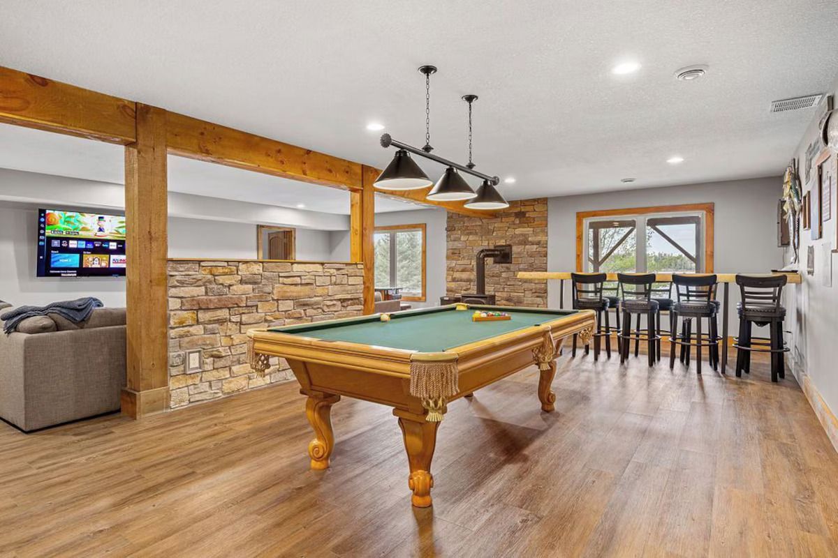 Basement with pool table, bar seating, and TV on a stone accent wall. Wooden beams and flooring.