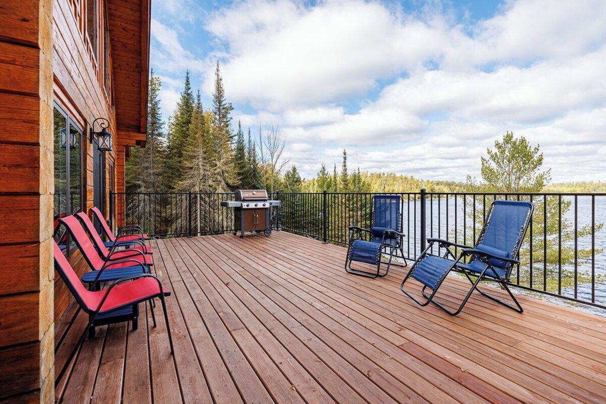 Wooden deck with lounge chairs, grill, and view of trees and lake under a cloudy sky.