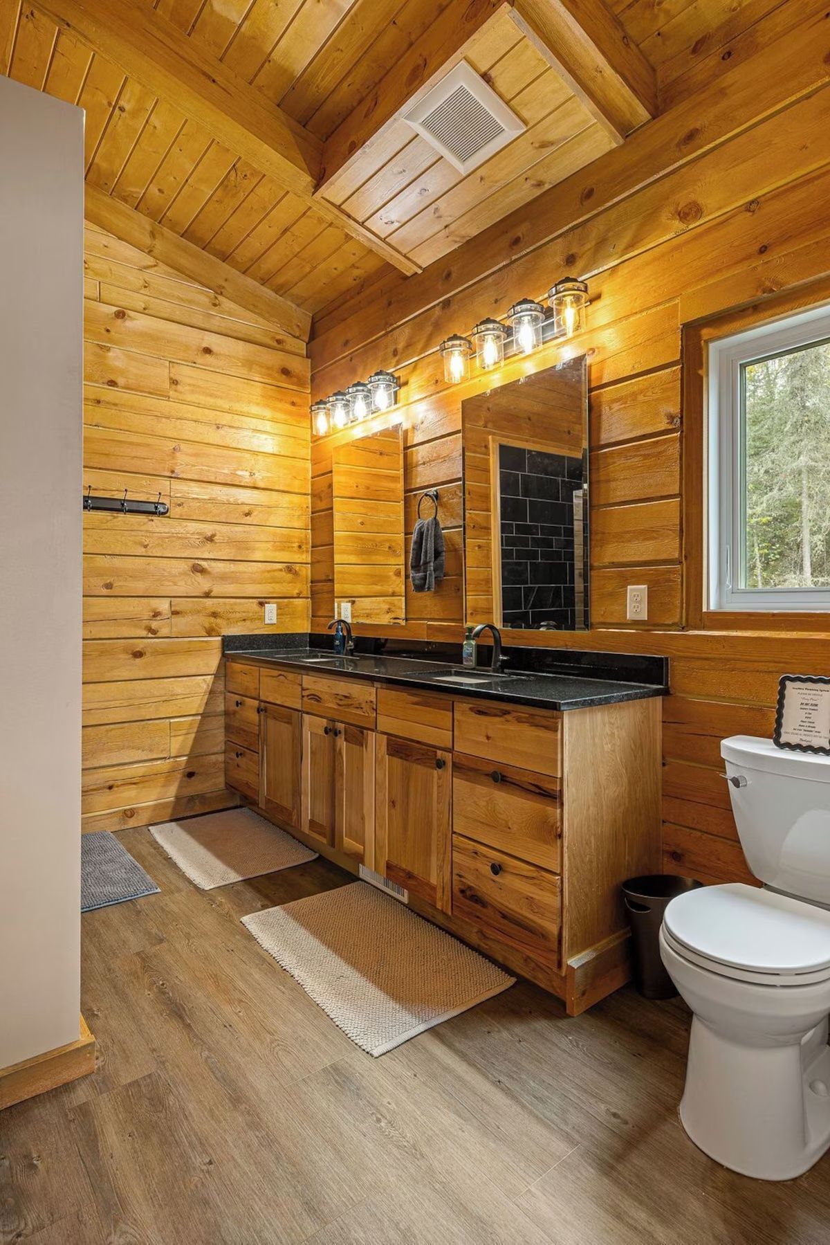 Rustic wood-paneled bathroom with a double vanity, black countertop, and a toilet.