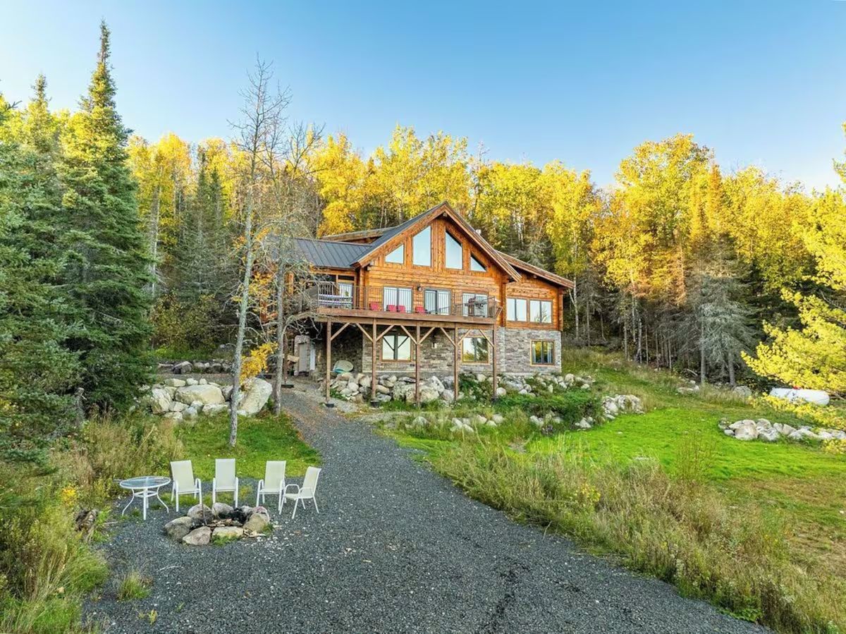 Rustic wooden cabin on a hill, with gravel driveway, chairs by fire pit, and trees in background.