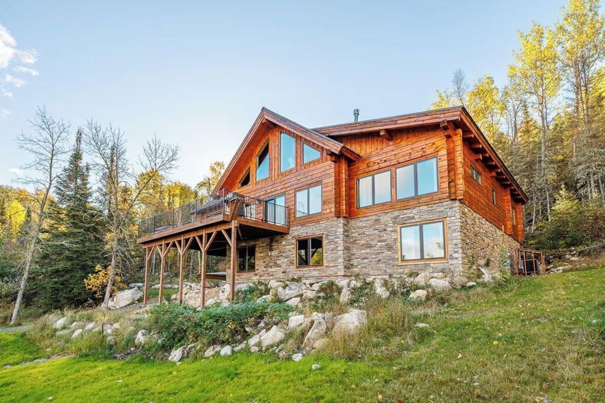 Two-story log cabin with stone base, wooden porch and balcony, set on a grassy hill surrounded by trees.