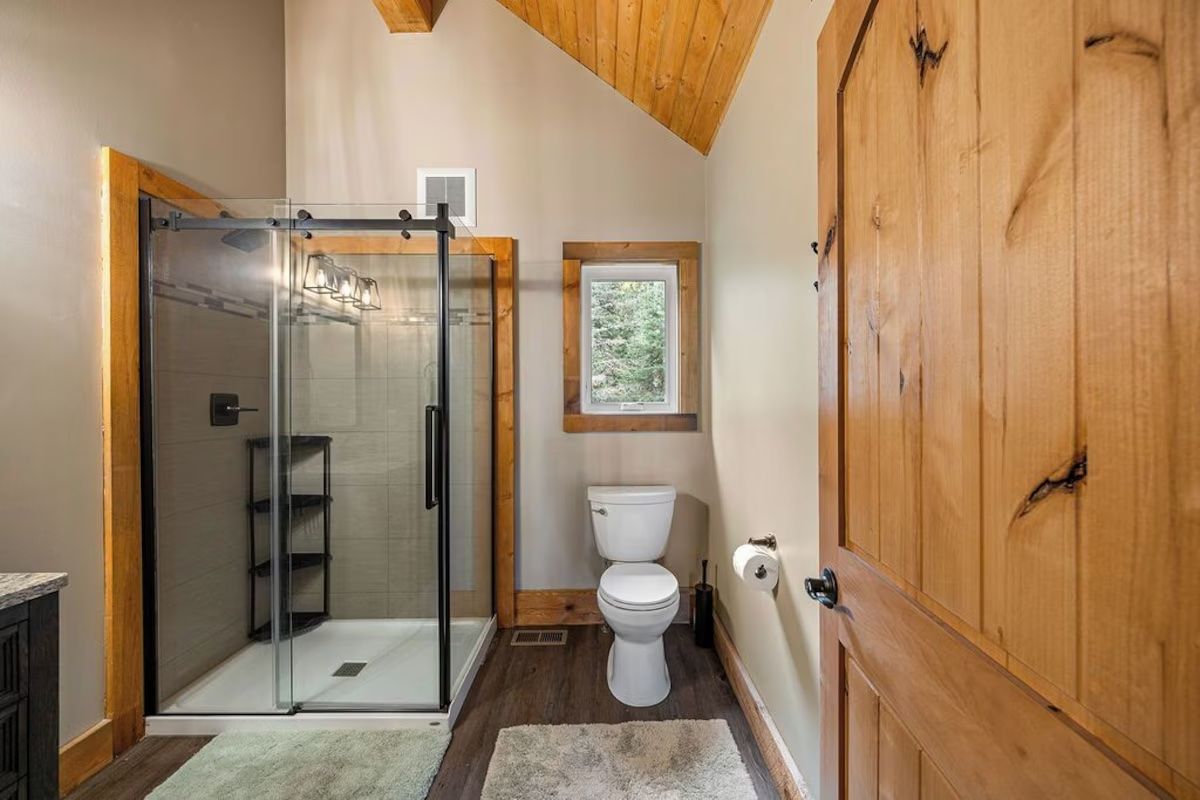 Bathroom with a glass shower, wooden door, toilet, and a small window.