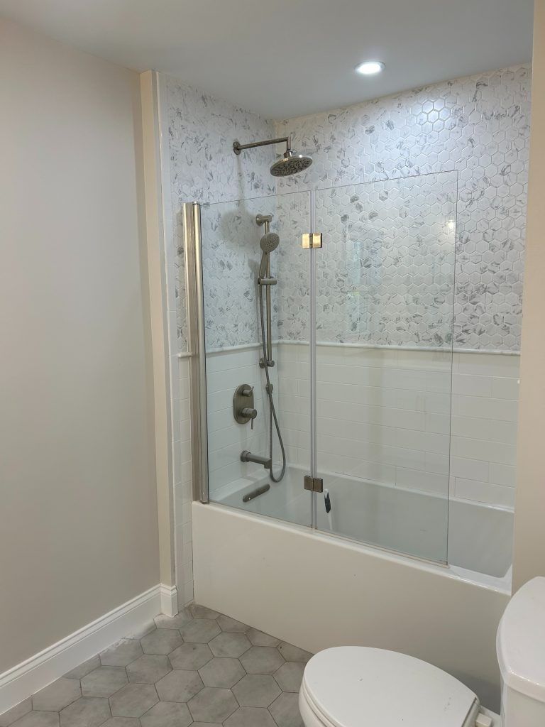 Shower stall with glass door, white tub, mosaic tile backsplash, silver showerhead, and a toilet.