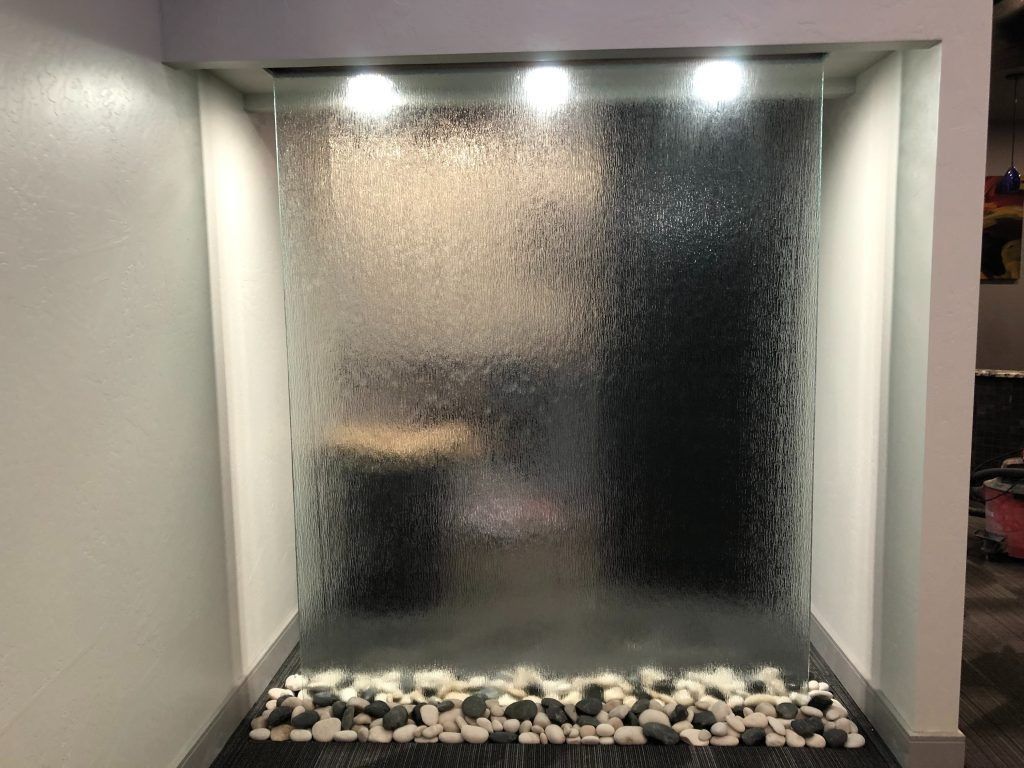 Glass waterfall feature with lights, black and white stones at the base, and a gray wall backdrop.