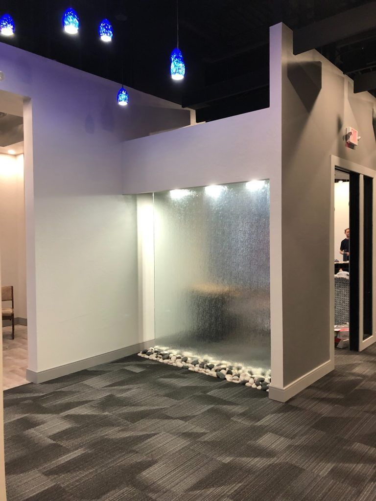Indoor water feature with cascading water on glass, rocks at base, and blue lights above.