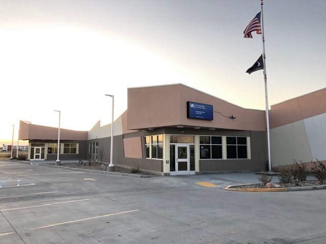 Tan and gray building with a blue sign, American flag, and parking area in front.