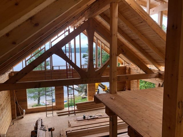 Interior view of a wooden house under construction; exposed beams and a lake visible through the window.