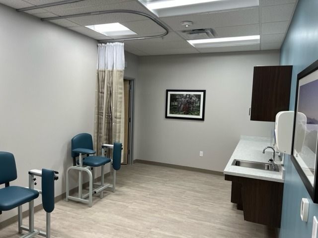 Examination room with blue chairs, sink, and privacy curtain. Neutral walls and floor.