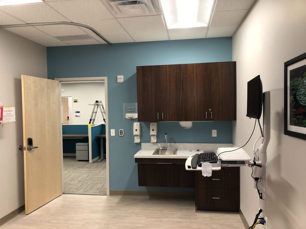 Medical exam room with sink, cabinets, monitor, and open doorway to another space.