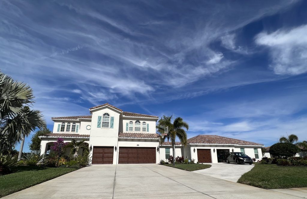 Large stucco home with brown garage doors and a long driveway under a blue sky with streaky clouds.