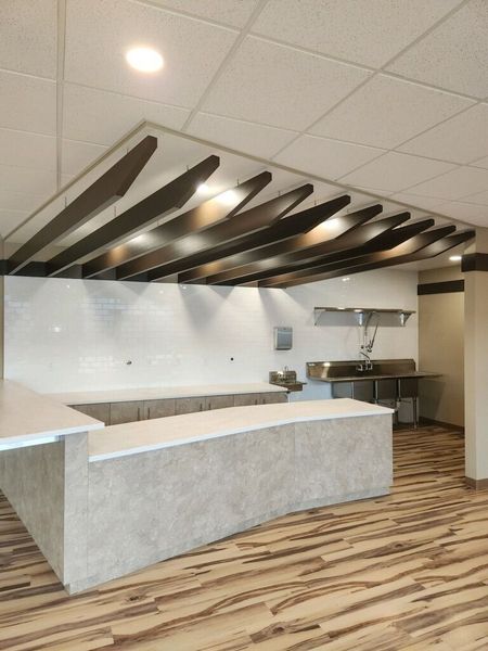 Modern commercial kitchen with a light-colored countertop and a wood beam ceiling. Stainless steel sinks are visible in the background.