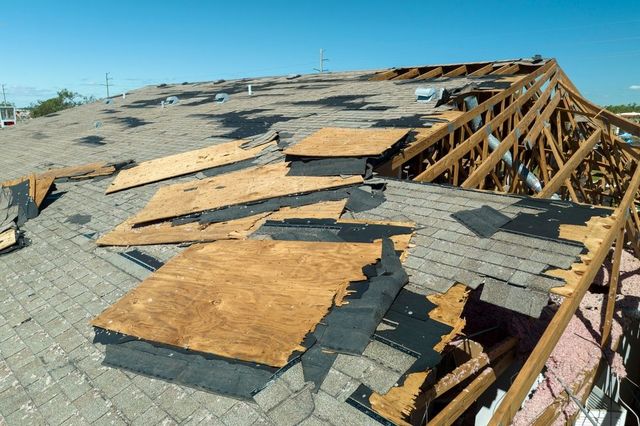 Damaged roof with missing shingles and exposed wooden beams under a bright blue sky.