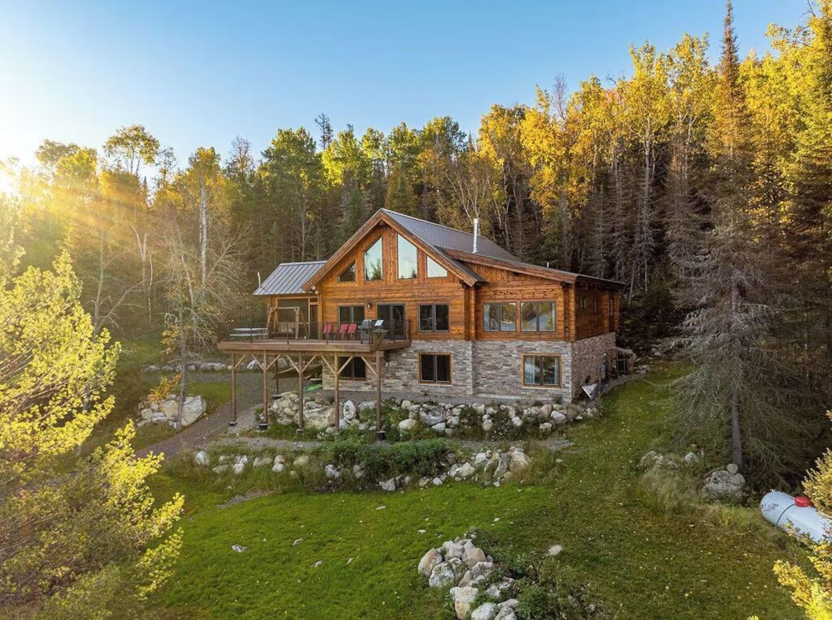 Log cabin nestled in a forest, deck, stone foundation, surrounded by trees and green grass.
