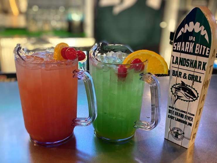 Two colorful tropical cocktails with fruit garnish beside a Shark Bites Landshark Bar & Grill menu tent